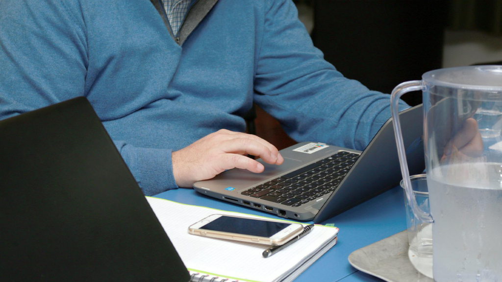 homem com a função de liderar a distância mexendo num notebook enquanto tem seu celular e um caderno de anotações sobre a mesa, este home veste um casaco azul